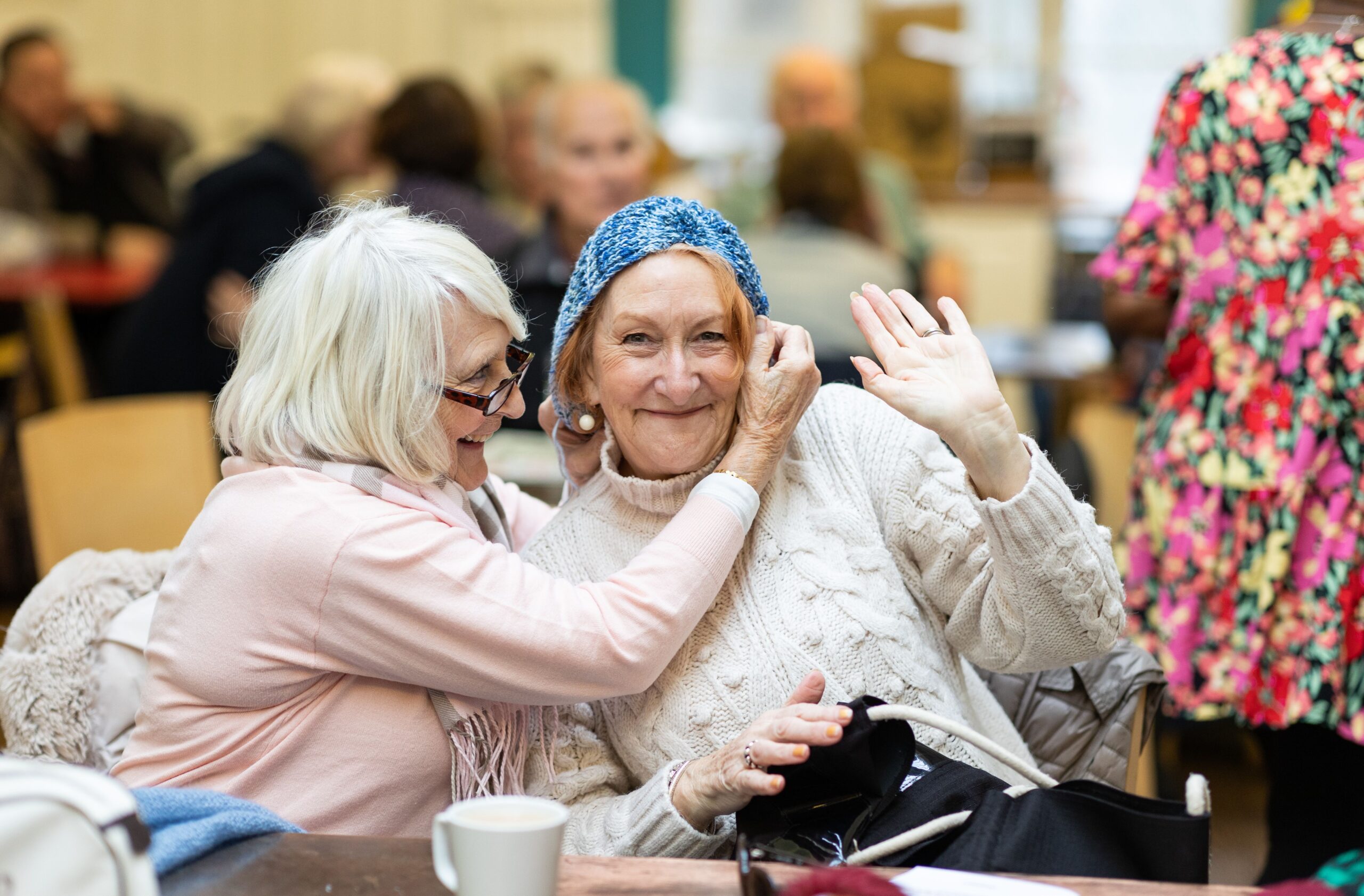 Two older women enjoying a friendly moment at an indoor social gathering, with one smiling and waving while wearing a blue knitted hat.